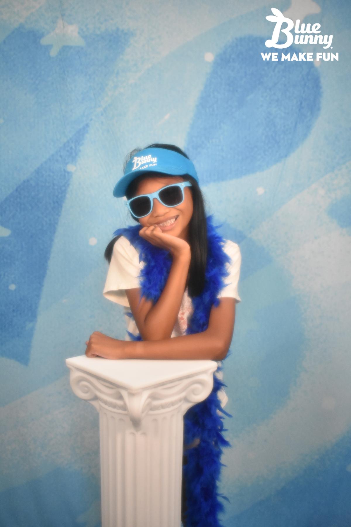 Girl leaning on a pillar holding her chin on her hand with sunglasses, a visor, and boa